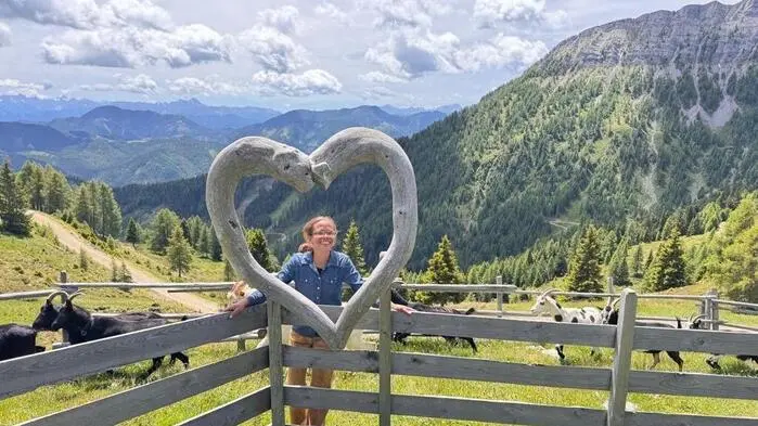 Eine Frau hinter einem Herz aus Holz, sie lächelt in die Kamera, hinter ihr Bergkulisse