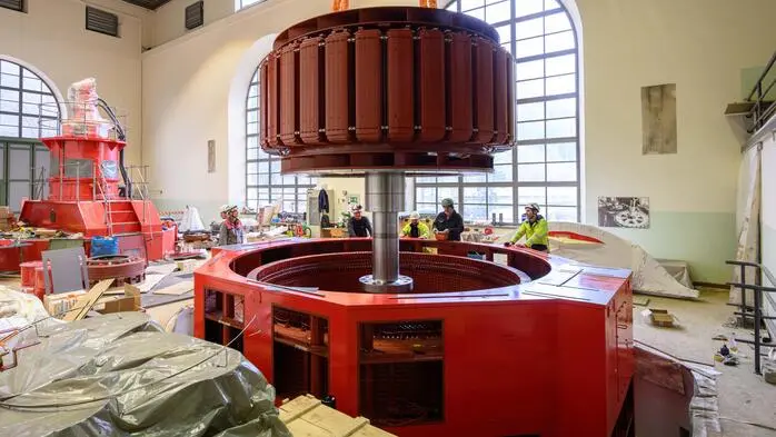 Ein Pkw-großer Generator wird im Krafthaus per Kranseil in seine runde Umhäusung gehoben, an deren Rand Arbeiter stehen