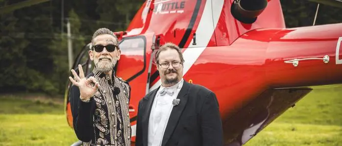 Ein Mann im extravaganten Outfit steht vor einem Helikopter. Neben ihm sein Butler in einem Frack. Der Mann signalisiert „OK“ in Richtung Kamera.
