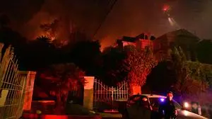 TOPSHOT - A police officer stands below burning brush at the Sunset Fire near Hollywood Blvd in the Hollywood Hills in Los Angeles, California, on January 8, 2025. At least five people have been killed in wildfires rampaging around Los Angeles, officials said on January 8, with firefighters overwhelmed by the speed and ferocity of multiple blazes -- including in Hollywood. (Photo by Robyn Beck / AFP)