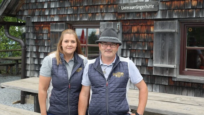 Eine junge Dame und ein Herr mit einem Filzhut stehen vor einer Almhütte