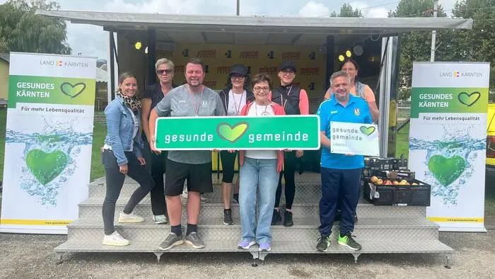 Eine Gruppe von Menschen steht im Freien und hält eine grüne Tafel in die Kamera. Darauf zu lesen ist „Gesunde Gemeinde“.