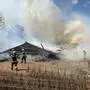Aufnahme eines niedergebrannten Hauses. Im Hintergrund steigt dichter Rauch auf. Es sind diverse Feuerwehrleute in Atemschutzausrüstung zu sehen