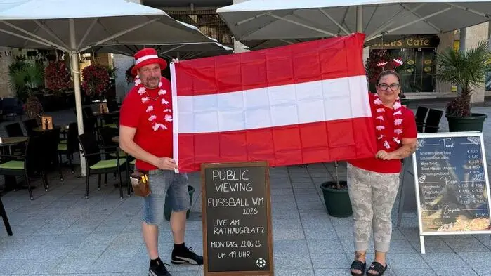 Eine Frau und ein Mann halten eine Österreichfahne in die Kamera, davor steht ein Schild, das das Public-Viewing ankündigt