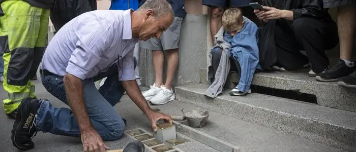 Ein Mann mit Hammer verlegt die Stolpersteine in den Boden und wird dabei von mehreren Umstehenden gefilmt