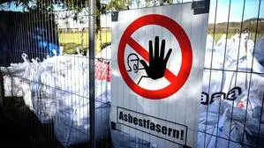 Große weiße Säcke hinter einem Bauzaun, darauf eine Tafel mit einer Warnung „Asbestfasern“