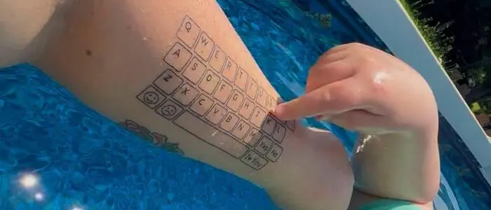 Ein Kind tippt mit dem Finger auf einen Unterarm, auf dem eine Tastatur tätowiert ist. Er befindet sich dabei in einem Pool.