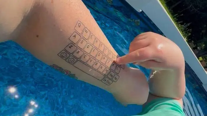 Ein Kind tippt mit dem Finger auf einen Unterarm, auf dem eine Tastatur tätowiert ist. Er befindet sich dabei in einem Pool.