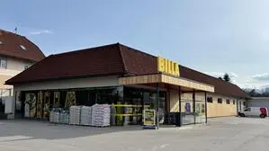 Blick von außen auf eine renovierte Billa-Filiale. Der Eingang und eine Seite sind mit Holz verkleidet, links vom Eingang stehen Säcke mit Erde zum Verkauf bereit