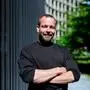 Stefan Bürscher ist Spitzenkandidat der Liste Gaza in Graz, er posiert mit verschränkten Armen, im Hintergrund ist ein Gebäude und ein Baum