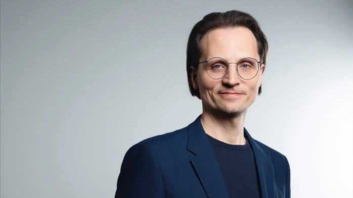 Ein Mann mit Brille, zurückgekämmten Haaren und schwarzer Hornbrille schaut mit einem leichten Lächeln in die Kamera 