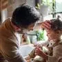 Father checking forehead of sick toddler daughter indoors in kitchen at home.