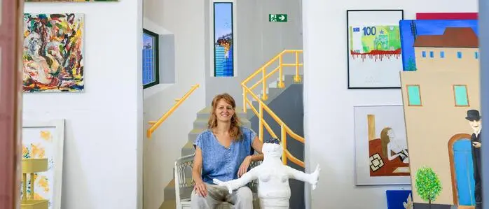 Eine Frau sitzt auf einem Stuhl und hält eine Puppe aus Gips. Im Hintergrund hängen jede Menge Bilder an der Wand.