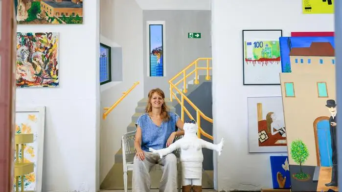 Eine Frau sitzt auf einem Stuhl und hält eine Puppe aus Gips. Im Hintergrund hängen jede Menge Bilder an der Wand.