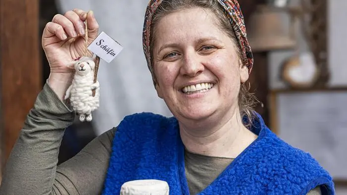 Eine Frau mit einem Spielzeugschaf und Käse in der Hand lacht in die Kamera