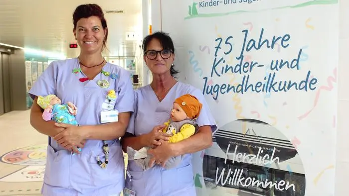 Zwei Pflegerinnen mit Puppen im Arm stehen vor einem Schild mit der Aufschrift „75 Jahre Kinder- und Jugendheilkunde“