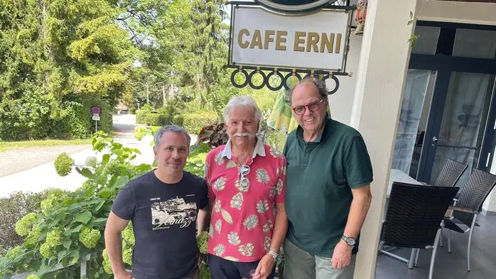 Drei Männer posieren für ein Foto und lächeln in die Kamera. Im Hintergrund sieht man ein Schild mit der Aufschrift „Café Erni“.