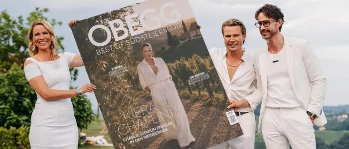 Zwei Männer und eine Frau, alle in Weiß gekleidet, präsentieren eine überdimensionale Magazin-Titelseite.