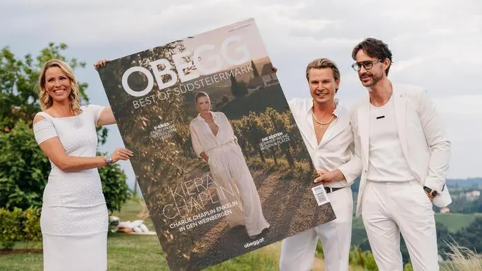 Zwei Männer und eine Frau, alle in Weiß gekleidet, präsentieren eine überdimensionale Magazin-Titelseite.