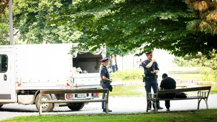 Zwei Beamte kontrollieren gerade einen Mann, der mit dem Rücken zur Kamera auf einer Bank im Park sitzt.