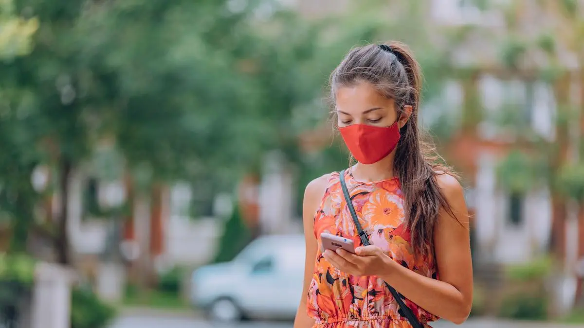 COVID-19 mask wear mandatory in city. Asian woman walking using mobile phone wearing face mask protection as prevention for coronavirus outside in city park summer lifestyle outdoor. Corona virus.