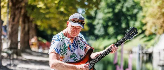 Gitarrist mit Schildkappe, Sonnenbrille und buntem Hemd sitzt mit E-Gitarre vor einem wasserführenden Kanal und Laubbäumen | Didi Baumgartner im Lendhafen in seiner Heimat Klagenfurt