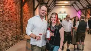 Ein Mann und eine Frau lachen in die Kamera. Der Mann hält eine Flasche Wein und ein Weinglas in seinen Händen.