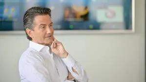 Auf dem Bild ist ORF-Generaldirektor Roland Weißmann zu sehen, der in einem modernen Konferenzraum sitzt. Er trägt ein weißes Hemd und hat dunkles, sorgfältig frisiertes Haar. Seine Körperhaltung ist entspannt, er hat einen Arm verschränkt und berührt mit der anderen Hand sein Kinn. Im Hintergrund befindet sich ein großer Bildschirm, auf dem unscharf verschiedene Symbole und Inhalte zu erkennen sind. | ORF-Generaldirektor Roland Weißmann im Interview