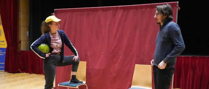 Eine Frau und ein Mann stehen vor einem roten Vorhang, die Frau hat einen Fuß auf einem Sessel