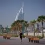 Das Bild zeigt das Luxushotel Burj Al Arab in Dubai, Palmen und eine Promenade mit einem Läufer im Vordergrund. 