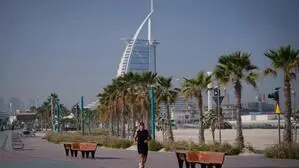 Das Bild zeigt das Luxushotel Burj Al Arab in Dubai, Palmen und eine Promenade mit einem Läufer im Vordergrund. 