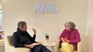 Zwei Frauen sitzen auf hellen Stühlen, dazwischen steht ein kleiner Tisch mit Blumen darauf