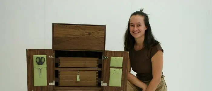 Eine junge Frau mit langen, braunen Haaren kniet vor ihrem Meisterstück, einer Holzkommode für Schneiderinnen und Schneider