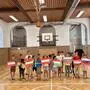 Mehrere Volksschulkinder stehen in einem Turnsaal und halten selbstgestaltete Länderflaggen in die Höhe