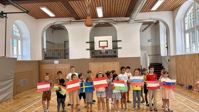 Mehrere Volksschulkinder stehen in einem Turnsaal und halten selbstgestaltete Länderflaggen in die Höhe