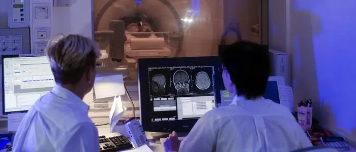 Zwei Ärztinnen sitzen vor einem Bildschirm, im Hintergrund wird eine Computertomographie einer Person gemacht.