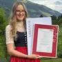 Eine junge Frau in Tracht steht auf einer Wiese und hält eine Urkunde in die Kamera