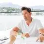 Ein Mann im weißem Poloshirt sitzt an einem Tisch am See und spricht