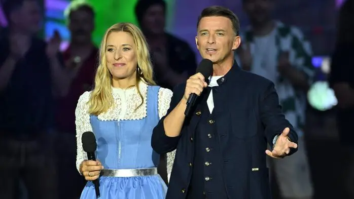 Ein Mann und eine Frau moderieren auf einer Bühne. Beide halten ein Mikrofon in ihren Händen.