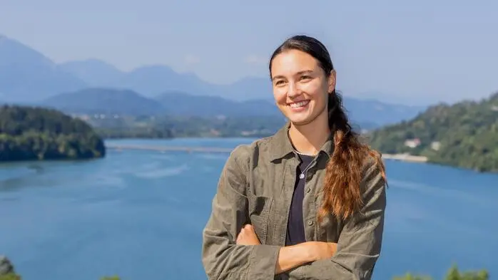 Eine braunhaarige Frau mit Zopf lacht in die Kamera, im Hintergrund Wasser und Berge