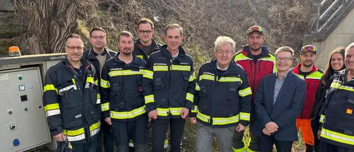 Eine Gruppe von Männern in Feuerwehruniform steht vor einem Stromkasten und lächelt in die Kamera.