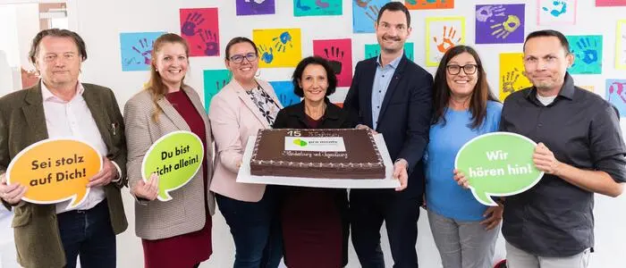 Ein Gruppenfoto mit verschiedenen Personen in förmlicher Kleidung. In Hintergrund sind bunte Papiere an der Wand. Die Frau in der Mitte hält eine große Schokotorte.