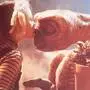 Das Bild zeigt die berühmte Szene aus dem Film „E.T. - Der Außerirdische“, zwischen dem kleinen Mädchen Gertie, gespielt von Drew Barrymore, und dem außerirdischen Wesen E.T.. Das Mädchen trägt blonde Zöpfe und ein gestreiftes Langarmshirt und eine Latzhose. Es beugt sich vor und gibt dem Außerirdischen einen sanften Kuss auf die Nase. Das Wesen hat eine braune, faltige Haut, einen großen Kopf und große Augen. Es hält einen Blumentopf mit gelben Blumen in den Händen. | E.T. - Der Außerirdische gehört zu den Filmklassikern.