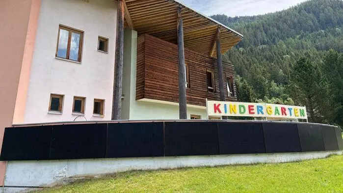 Foto eines hellroten Gebäudes, eingebettet in eine Waldlandschaft. Zu sehen ist der Schriftzug „Kindergarten“, darunter ist eine Wand voller PV-Paneele.