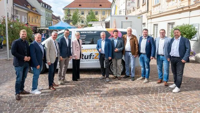 Verschiedene Männer in Anzügen und eine Frau stehen vor einem weißen Taxibus. Sie sind umgeben von einer mitteleuropäischen Altstadt. Der Himmel ist leicht bewölkt.