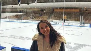 Eine dunkelhaarige, junge Frau mit dicker Jacke lacht in die Kamera und steht vor einem Eishockey-Feld