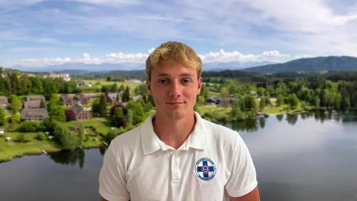 Porträt eines jungen Mannes in einem Wasserrettungspoloshirt. Im Hintergrund sieht man eine Drohnenaufnahme von einem See