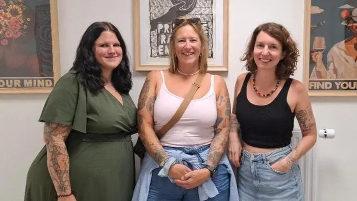 Drei Frauen stehen vor einer Wand mit bunten Bildern und lächeln in die Kamera