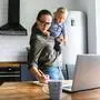 Busy mom works at home with an active toddler on her arms. Mother in glasses is watching on laptop and writing notes in the kitchen while holding kid