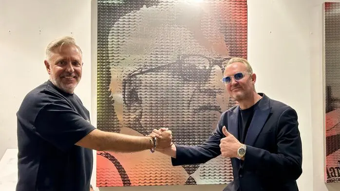 Zwei Männer stehen in schwarzer Kleidung vor einem Kunstwerk, das Enzo Ferrari zeigt und schlagen ihre Hände ein
